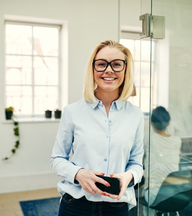 Confident young businesswoman wearing glasses standing with her cellphone in an office with colleagues at work in the background